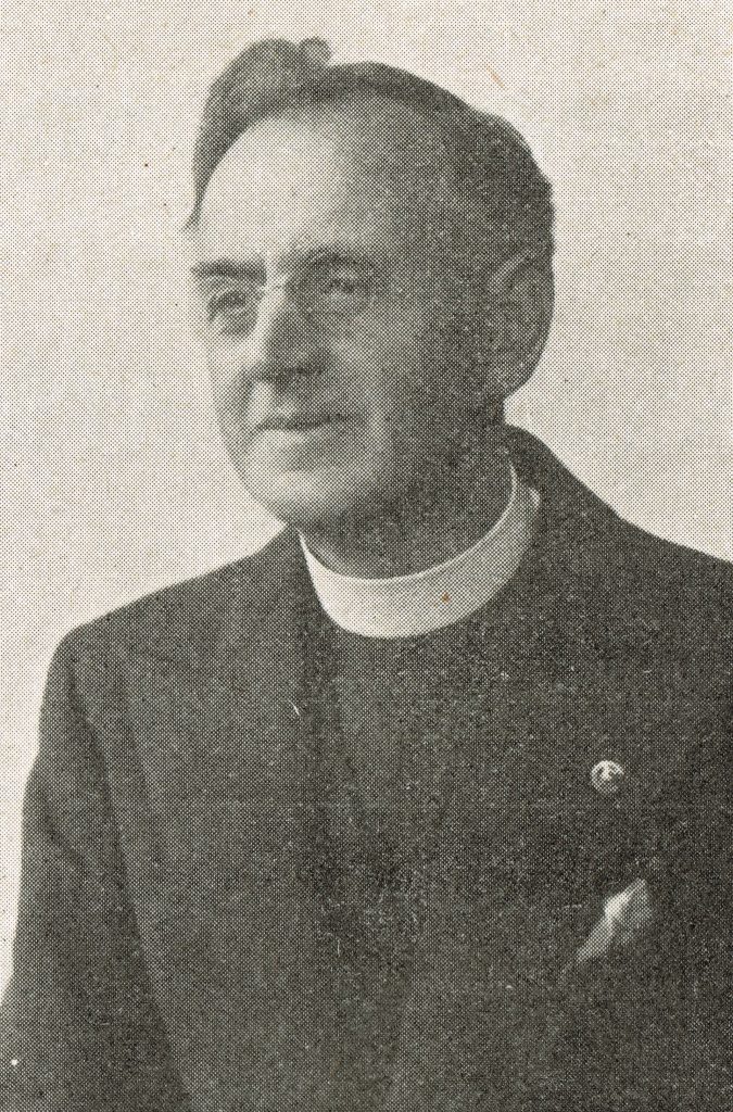 Portrait photograph. Jesse is clean shaven and wears glasses and a wide full clerical collar