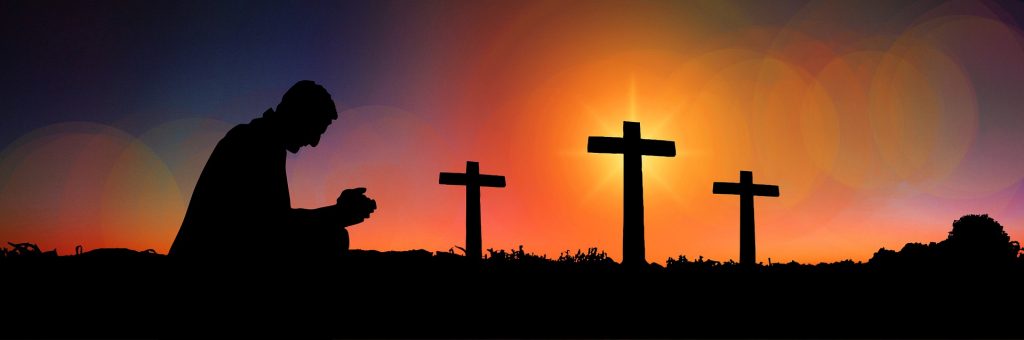 A  silhouetted figure kneels in prayer in front of three crosses outlined by the setting sun.