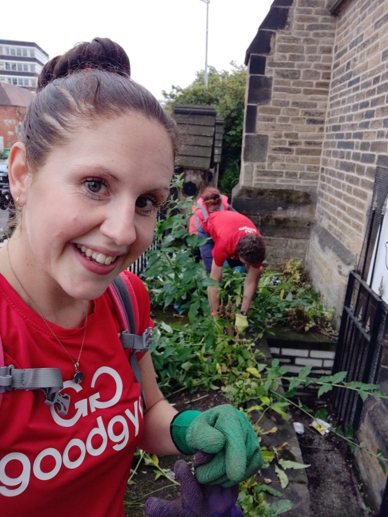 Sian holds some weeds in her hands as behind her two others are busy removing weeds from the front of the church