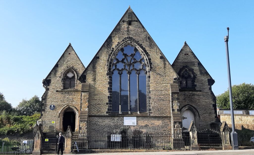 View of the front of Wycliffe Congregational Church from the other side of Wellington Road North. The left hand side door is open and a bicycle is fastened to the railings. A man is just turning to enter the gate on a bright sunny day.