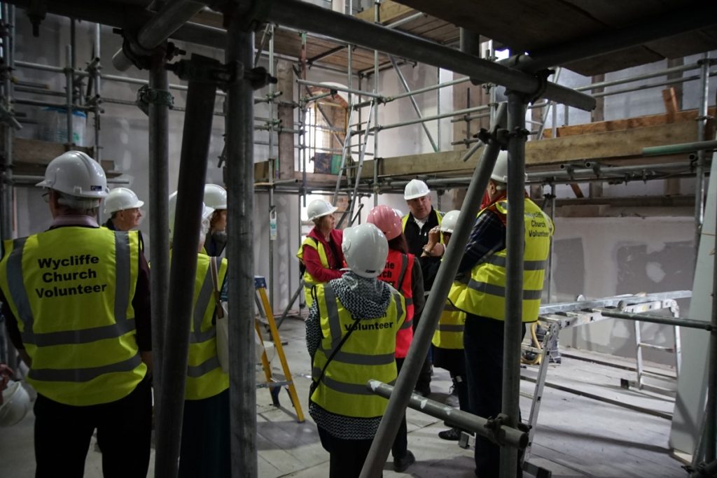 Visitors in hard hats stand amongst scaffolding listening to a speaker