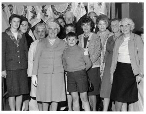 A group of ladies stand in front of hanging embroidery with a young boy in the forefront