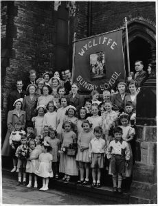 A group of adults and children stand with the Sunday School banner at the North East entrance