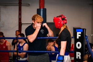 A trainer holds his fists up demonstrating moves to a girl in protective gear in the ring