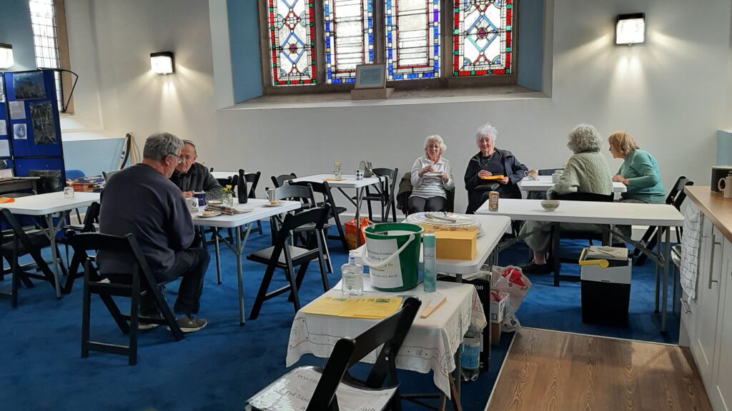 A view of the blue carpeted community room in Wycliffe with ladies enjoying a cup of tea and cake, smiling. On the right, two gentlemen are deep in conversation
